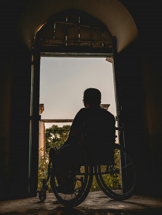 uma pessoa dentro de uma casa e sentada em cadeira de rodas olha para fora por uma porta aberta. A imagem interna da casa está escura (contraluz) e do lado de fora há luz que faz vê céu azul e alguma árvores e mato.